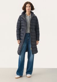 Marineblauwe pufferjas met een capuchon, gouden hardwareaccenten, lange lengte en gestikte delen. Gecombineerd met een grijze gebreide cardigan en wijde broeken.