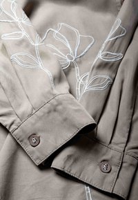 Tissu beige avec broderie florale blanche et poignets pliés attachés par des boutons marron sur un vêtement, montré en gros plan.