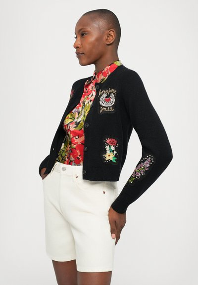 Woman with short hair wearing a black cardigan with floral patches over a red floral blouse and white high-waisted shorts, hands in pockets.