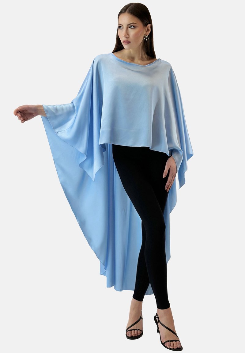 Ljusblå cape-top i mjukt tyg, med vida ärmar och en flödande silhuett, kombinerad med svarta leggings och sandaletter.