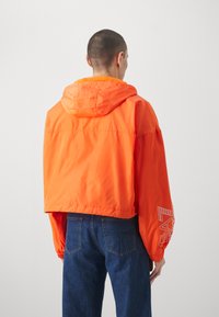 Kort orange vindtæt jakke med hætte, der har elastiske manchetter og et grafisk print på venstre ærme. Båret over blå denim jeans.