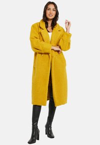 Abrigo amarillo de gran tamaño hecho de una tela texturizada, con cuello de chal, dos bolsillos frontales y botones dobles.