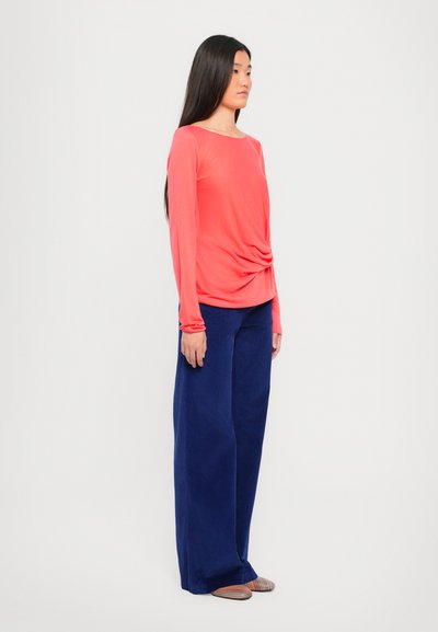Top a maniche lunghe corallo con nodo drappeggiato in vita, abbinato a pantaloni larghi blu navy e ballerine a strisce multicolori.