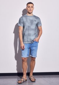 Šedé tričko s krátkým rukávem a vzorem, spárované s světle modrými džínovými šortkami. Muž stojí oblečený v hnědých sandálech, s rukama v kapsách, před hladkou zdí.