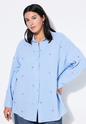 Frau mit dunklen Haaren trägt hellblaues Hemd mit lila Herzmustern, posiert mit einer Hand in der Hüfte vor weißem Hintergrund.