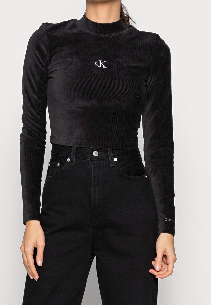 Svart kordfloret crop top med långa ärmar, hög halsringning och broderad "CK"-logotyp. Tillsammans med högmidjade svarta jeans.