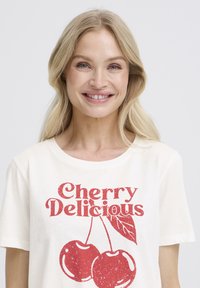 T-shirt crème clair avec imprimé "Cherry Delicious" en rouge usé, illustré de deux cerises et d'une feuille. Matière coton douce, coupe décontractée.