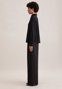 Blusa nera con colletto, maniche lunghe e polsini con bottoni abbinata a pantaloni neri a vita alta. Tessuto liscio con taglio sartoriale.