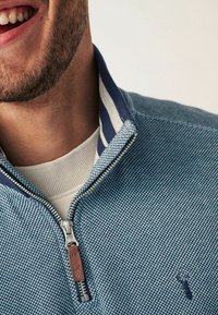 Pull-over zippé bleu en tissu tricoté texturé, doté d'un col rayé et d'un curseur de fermeture éclair en cuir marron avec un détail logo.