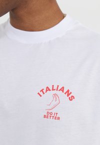 Balta medvilninė marškinė su raudona grafika, kurioje pavaizduotas rankos gestas, o virš ir žemiau vaizdo yra tekstas "ITALAI DARO GERIAU".