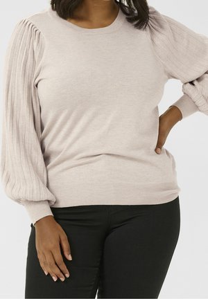 Femme portant un pull beige clair avec des manches longues plissées et un pantalon noir, une main sur la hanche et l'autre reposant sur le côté.