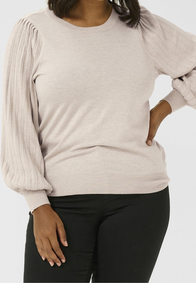 Femme portant un pull beige clair avec des manches longues plissées et un pantalon noir, une main sur la hanche et l'autre reposant sur le côté.