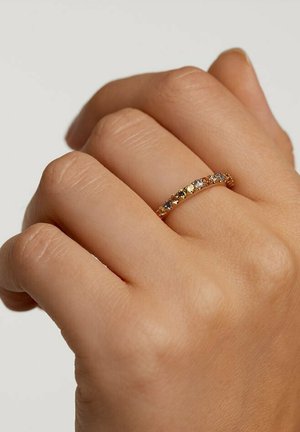 Anello d'oro con gemme multicolori alternate incastonate in una fascia curva, appoggiato su una mano con texture della pelle visibile.