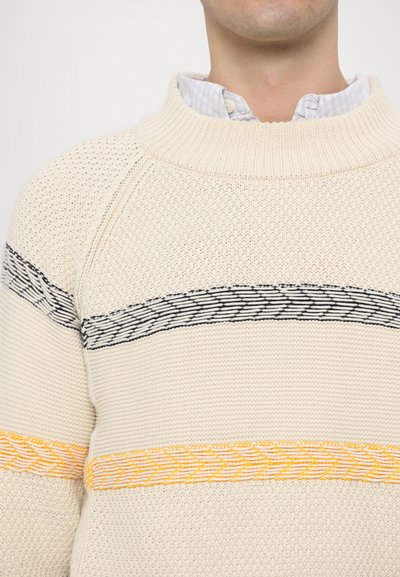 Pull crème en tricot avec col rond. Présente des motifs rayés horizontaux noirs et orange ainsi qu'un tissu tissé texturé.