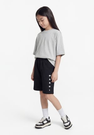 Dieťa má na sebe sivé oversized tričko, čierne krátke nohavice s vertikálnym nápisom „MARNI“, biele ponožky a čierno-biele tenisky, stojí na bielom pozadí.