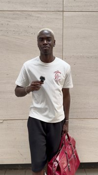 T-shirt bianco a maniche corte con grafica rossa, pantaloni corti neri, che tiene un microfono nero e una grande borsa di pelle rossa con accenti argentati.