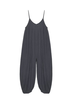 Femme debout contre un mur blanc portant une combinaison ample sans manches gris foncé avec chevilles froncées et des sandales à plateforme noires.