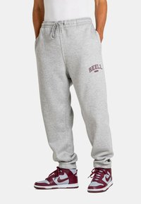 Graue Baumwoll-Sweatpants mit verstellbarem Bund und seitlichen Taschen, mit einem bordeauxfarbenen "REELL"-Logo. Kombiniert mit bordeauxfarbenen und grauen High-Top-Sneakern.