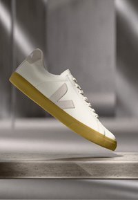 Hvite joggesko med glatt skinn-overdel, beige detaljer og en teksturert gul gummisåle. Har en distinkt logo på siden.