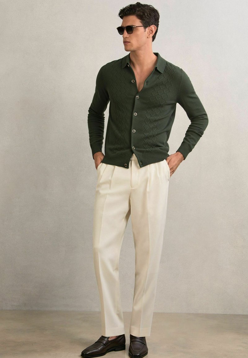 Cardigan verde tricotat cu model în diamant, cu nasturi în față, asortat cu pantaloni cremoși plisati și pantofi negri slip-on. Ochelari de soare purtați.