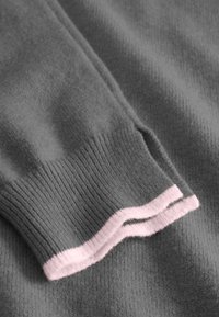 Grauer Pulloverstoff mit gerippter Textur und einem rosa Akzent am Bündchen. Weiches und glattes Material mit einem einfachen, aber klassischen Design.