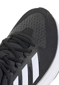 Gros plan d'une chaussure de sport noire avec des lacets noirs, des bandes latérales blanches et une semelle blanche texturée.
