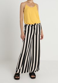 Top sin mangas amarillo combinado con pantalones de pierna ancha a rayas en blanco y negro, que cuentan con cintura con cordón y sandalias de tiras trenzadas.