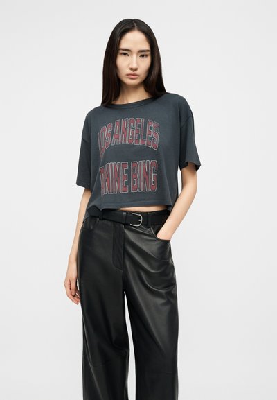 Hosszú sötét hajú nő, laza, sötétszürke, rövid felsőt visel, amelyen a „LOS ANGELES NINE BING” felirat szerepel, valamint fekete, széles szabású bőr nadrágot.