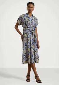 Moteris vilkinti mėlyną ir geltoną gėlių midi suknelę su trumpomis rankovėmis, juosmens diržu ir juodais aukštakulniais sandalais.