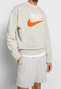 Ljusgrå sweatshirt med rundad hals och orange Nike-logotyp, med ribbade muddkanter, avslappnad passform, bärs över en vit undertröja och rutiga grå shorts.