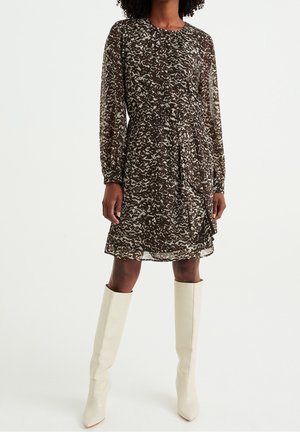 Femme portant une robe marron et blanche à motifs abstraits, manches longues, longueur genoux, avec des bottes blanches montantes sur fond blanc.