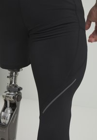 Svarta atletshorts med en figursydd design, med en reflekterande grå söm. En protesben syns under shortsen.