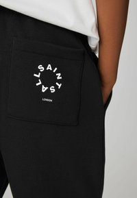 Sorte sweatpants med en baglomme og et cirkulært hvidt grafisk tekst, der står "SAINT LONDON." Stoffet er blødt og lidt tekstureret.