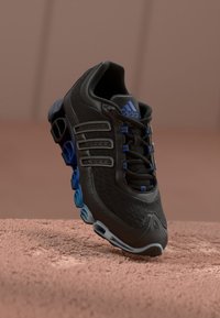 Black Adidas sneaker with blue accents, distinctive sole design, and three side stripes, displayed floating above a textured surface.