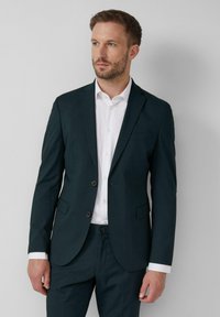 Blazer ajustado de un verde oscuro con dos botones, solapas notch y un bolsillo en el pecho. Llevado sobre una camisa blanca, combinado con pantalones a juego.