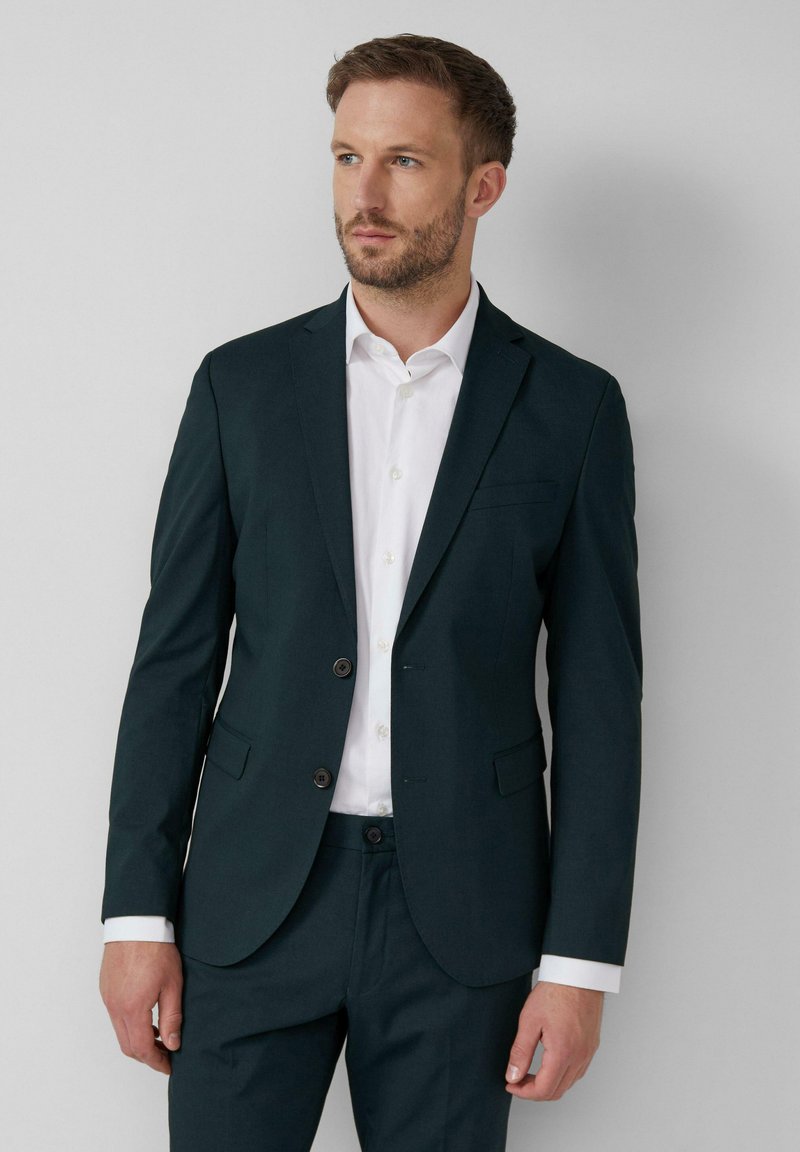 Blazer ajustado de un verde oscuro con dos botones, solapas notch y un bolsillo en el pecho. Llevado sobre una camisa blanca, combinado con pantalones a juego.