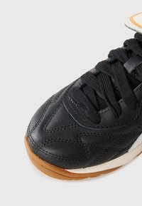 Zwarte sportieve schoen met een gestructureerde leren bovenkant, vetersluiting en een gumrubberen zool, voorzien van contrasterende lichte accenten aan de zijkant.