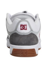 Bílé tenisky s šedými semišovými akcenty, červeným logem DC na patě a texturovanou gumovou podrážkou. Polstrovaný límec pro podporu.
