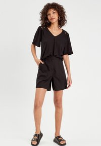 Schwarzes T-Shirt mit kurzen Ärmeln, strukturiertem Stoff und V-Ausschnitt; kombiniert mit schwarzen Shorts mit Fronttaschen. Sandalen mit zwei Riemen.