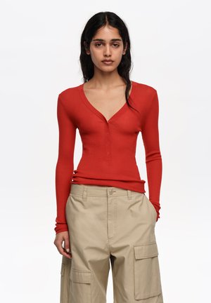 Ženska z dolgimi temnimi lasmi, oblečena v oprijet top z dolgimi r sleevesi rdeče barve in bež cargo hlače, stoji ob beli podlagi.