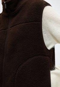 Gros plan d'un gilet en polaire texturé marron foncé avec une fermeture éclair à l'avant, porté sur un haut blanc en maille à manches longues.