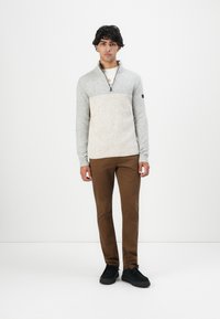 Jonge man staat in een tweekleurige grijze zip-up trui, bruine broek en zwarte schoenen voor een effen witte achtergrond.