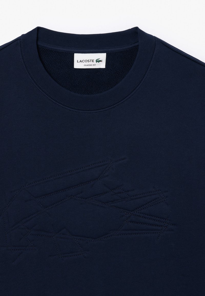 Granatowa bawełniana koszulka z teksturowanym, tonalnym logo krokodyla Lacoste. Klasyczny krój z okrągłym dekoltem i ribbowanym detalem kołnierza.