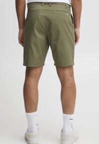 Férfi olíva zöld rövidnadrágban, fehér Nike zokniban és fehér sportcipőben áll, háttal egy sima fehér háttérnek.
