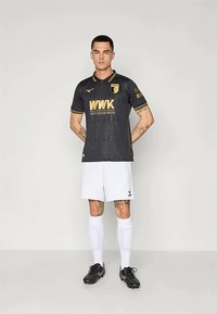 Μαλές ποδοσφαιριστής φορώντας μια μαύρη και χρυσή φανέλα της FC Augsburg, λευκά σορτς, λευκές κάλτσες μέχρι το γόνατο και μαύρα παπούτσια, στέκεται με τα χέρια πίσω από την πλάτη.