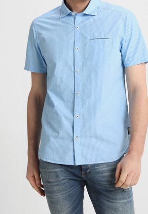 Mann trägt hellblaues kurzärmliges Hemd mit dezenten Streifen und blaue Jeans, steht vor einem einfarbigen Hintergrund.