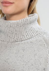 Hellgrauer Rollkragenpullover mit geripptem Kragen, der ein strukturiertes Strickmuster und verstreute schwarze Flecken aufweist. Nahaufnahme des Kragenbereichs.
