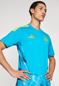 Young man with hands on hips wearing a bright blue sports jersey with yellow stripes and a team crest on the chest.