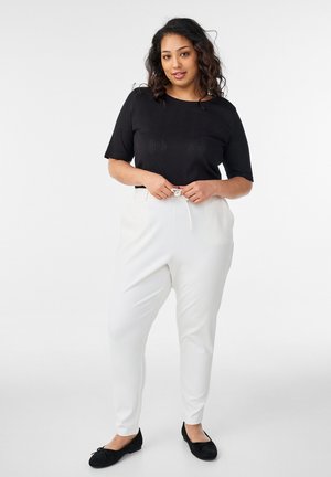 Kvinde binder snor på hvide taperede bukser, iført sort top med korte ærmer og sorte ballerinaer, stående mod hvid baggrund.