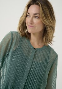 Blusa verde trasparente con pieghe testurizzate e piccoli motivi quadrati. Presenta un colletto rotondo e una chiusura con un unico bottone.